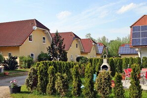 A row of yellow buildings with red tiled roofs, solar panels, and a well-maintained garden with hedges and a barbecue area. - Ferienhauser am Kummerower See, Verchen (Verchen)