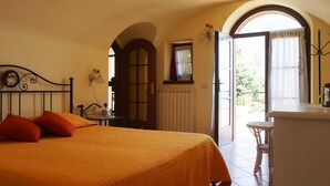 Ein Schlafzimmer mit einem Bett, einem kleinen Tisch, einem Sessel, einem Heizkörper und einer offenen Tür, die auf einen Balkon führt.