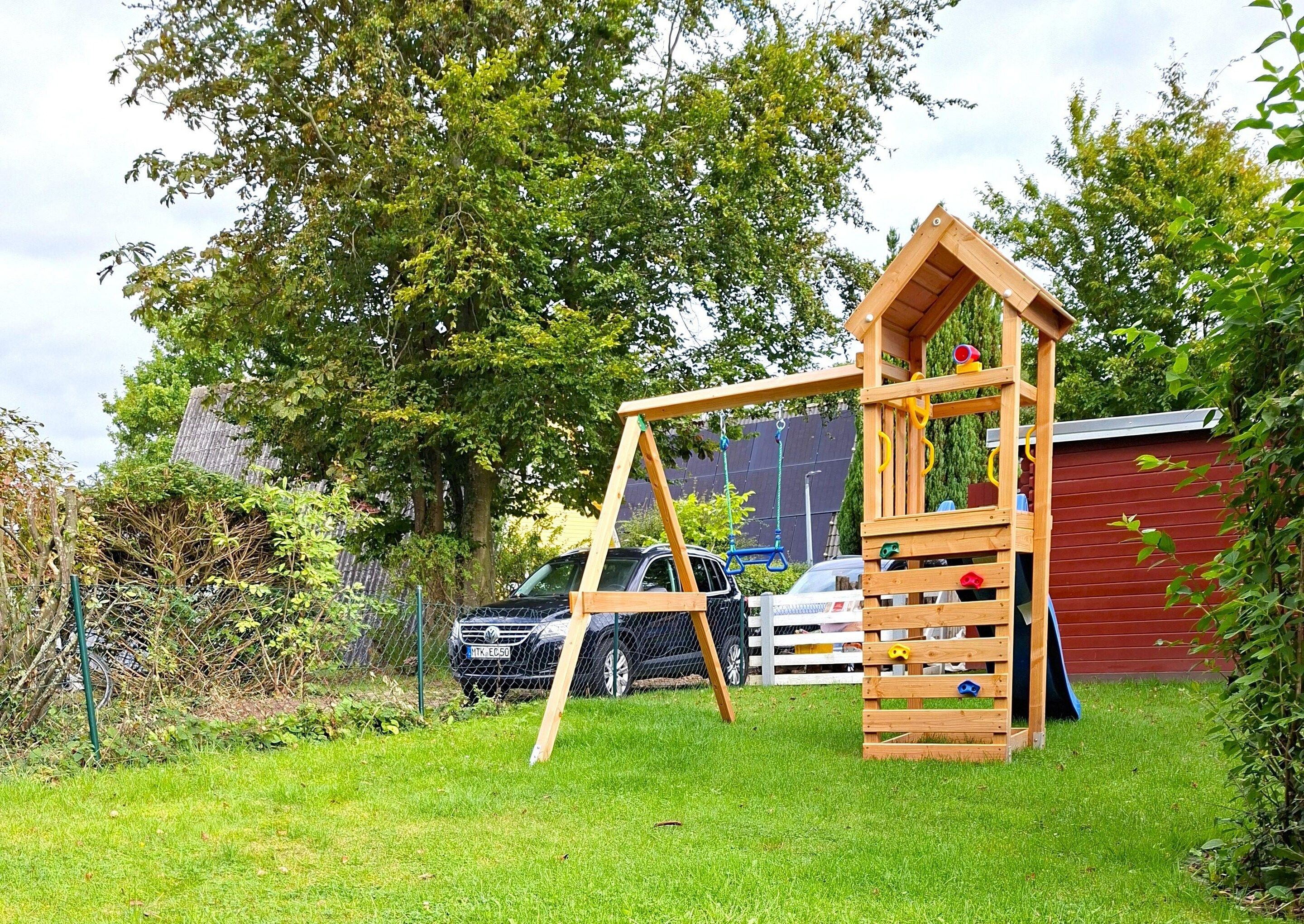 A wooden play structure in a grassy yard with a red building in the background.