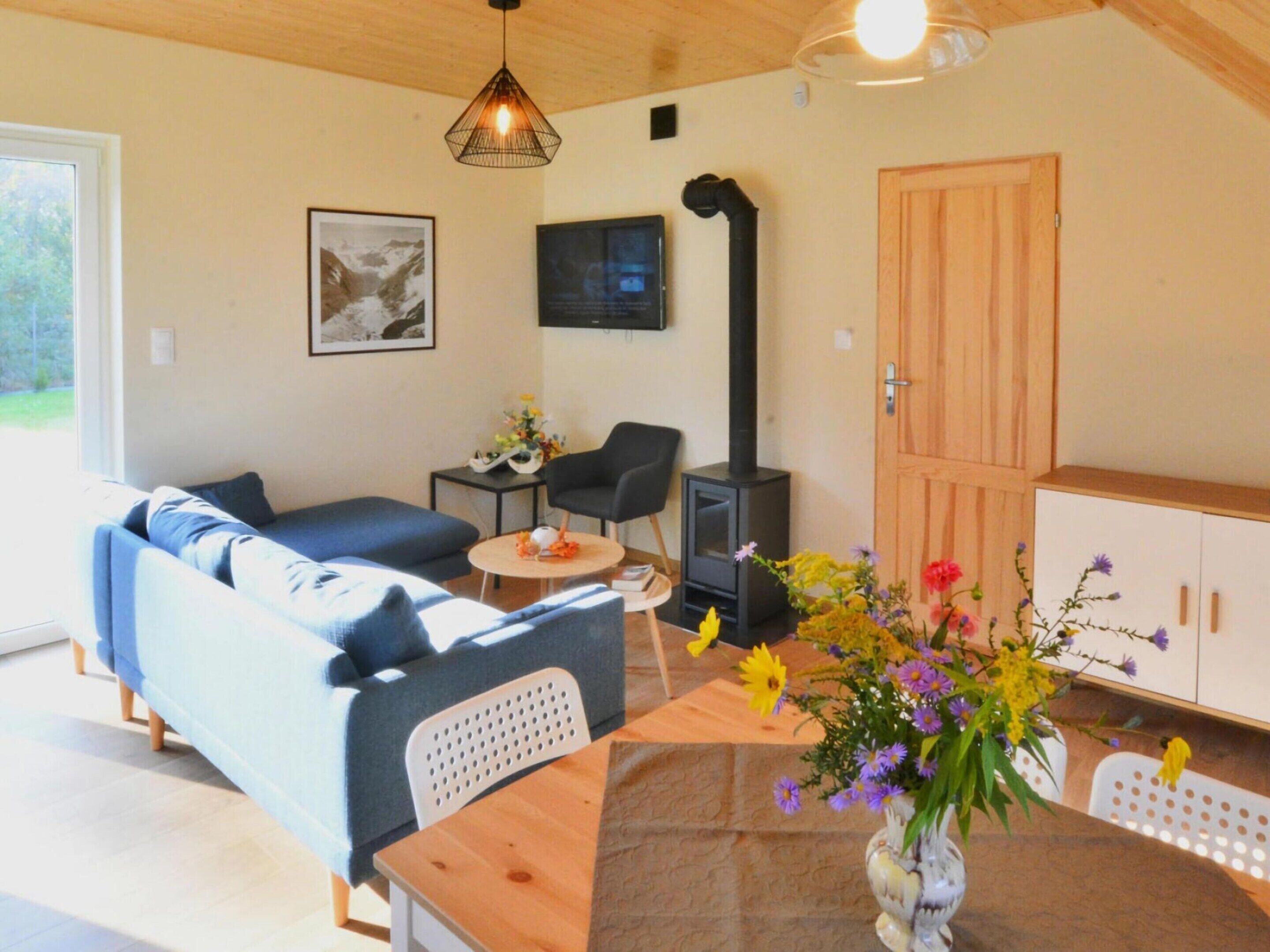 A cozy living room with a sofa, a table, a chair, a vase of flowers, a black stove, and a wooden cabinet.