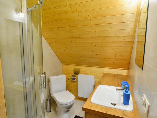 Une petite salle de bain aux murs lambrissés, dotée d’une douche en verre, de toilettes blanches et d’un meuble-lavabo en bois avec un évier et des distributeurs de savon bleus.