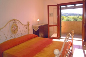 A bedroom with a bed, a nightstand, a dresser, a lamp, and an open door leading to a balcony with a view. - Apartment in Villaputzu With Garden (Domus de Maria)
