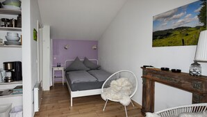 Quarto com cama, uma cadeira, prateleira com utensílios de cozinha e um quadro na parede.