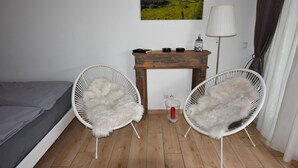 Quarto com duas cadeiras de vime branco, uma mesa de cabeceira de madeira com abajur, uma cama e um quadro de uma paisagem.