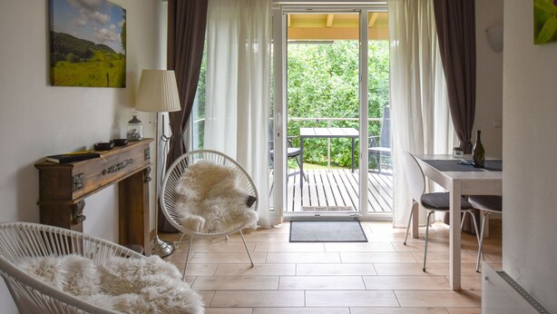 Quarto com escrivaninha de madeira, uma cadeira, poltrona confortável, luminária e vista para o verde através de uma porta de vidro.