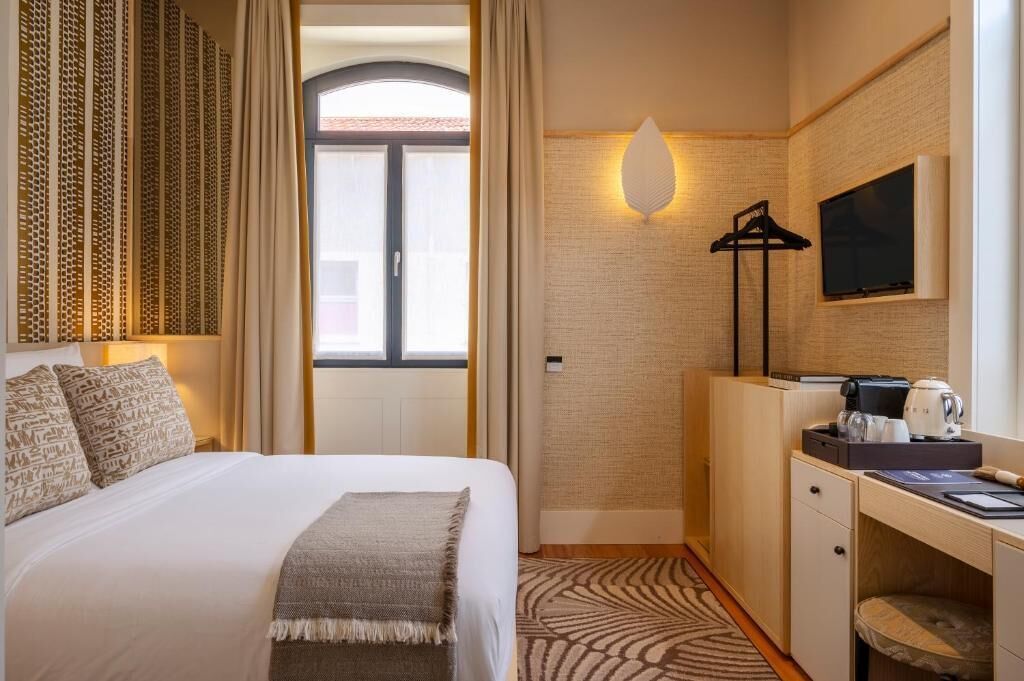 A hotel room with a bed, a desk with a coffee maker, a TV, and a window with curtains.