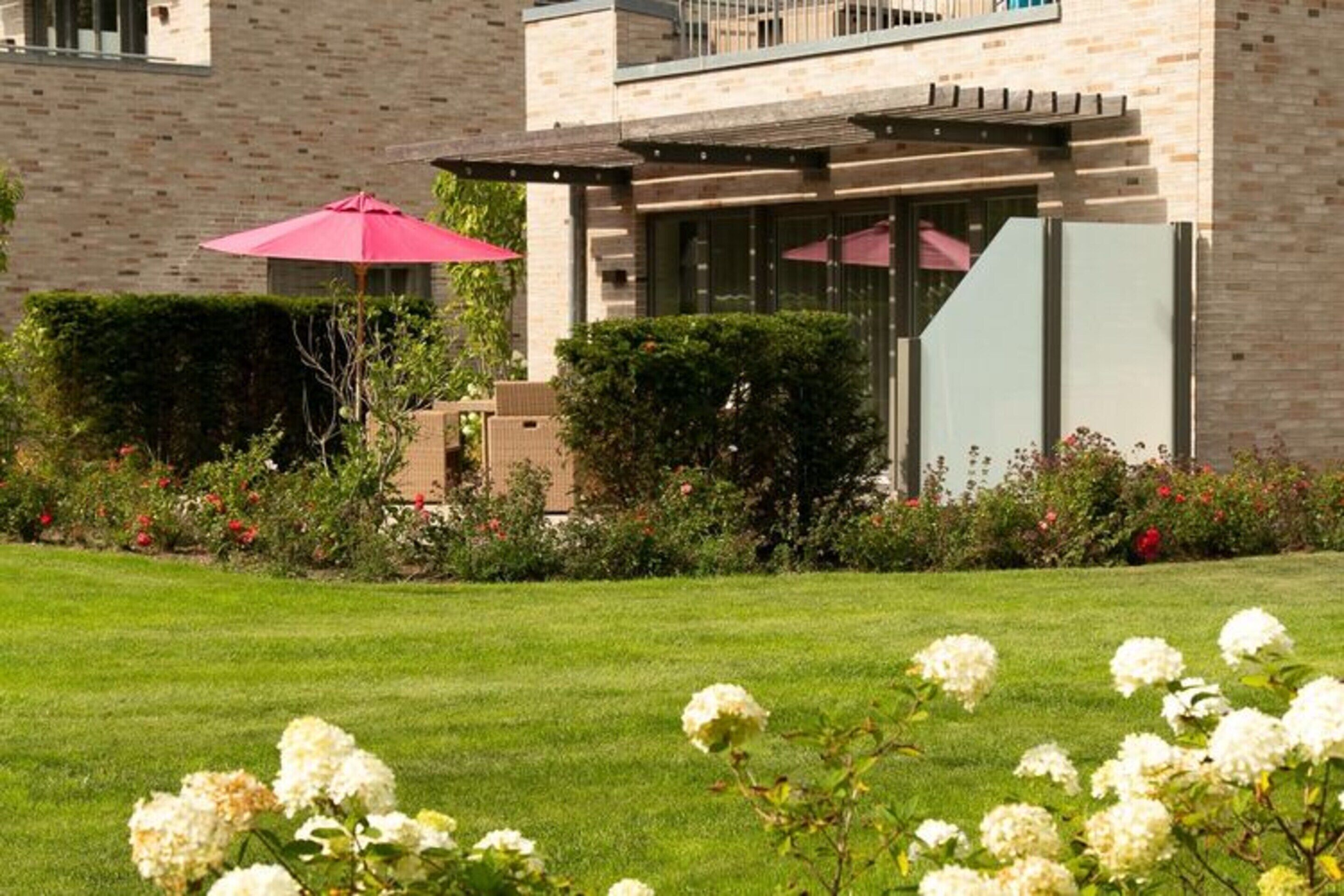 Un bâtiment moderne avec une façade en briques, un patio couvert et un jardin bien entretenu agrémenté de fleurs blanches.