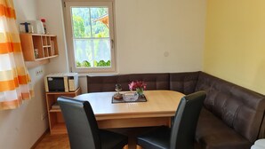 Un coin repas accueillant avec une table en bois, deux chaises noires et un banc en cuir marron. On aperçoit une petite étagère avec des livres et un four à micro-ondes.