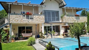 Une maison moderne à deux étages, dotée d’une piscine, d’une façade en pierre et d’un espace extérieur aménagé pour se détendre.