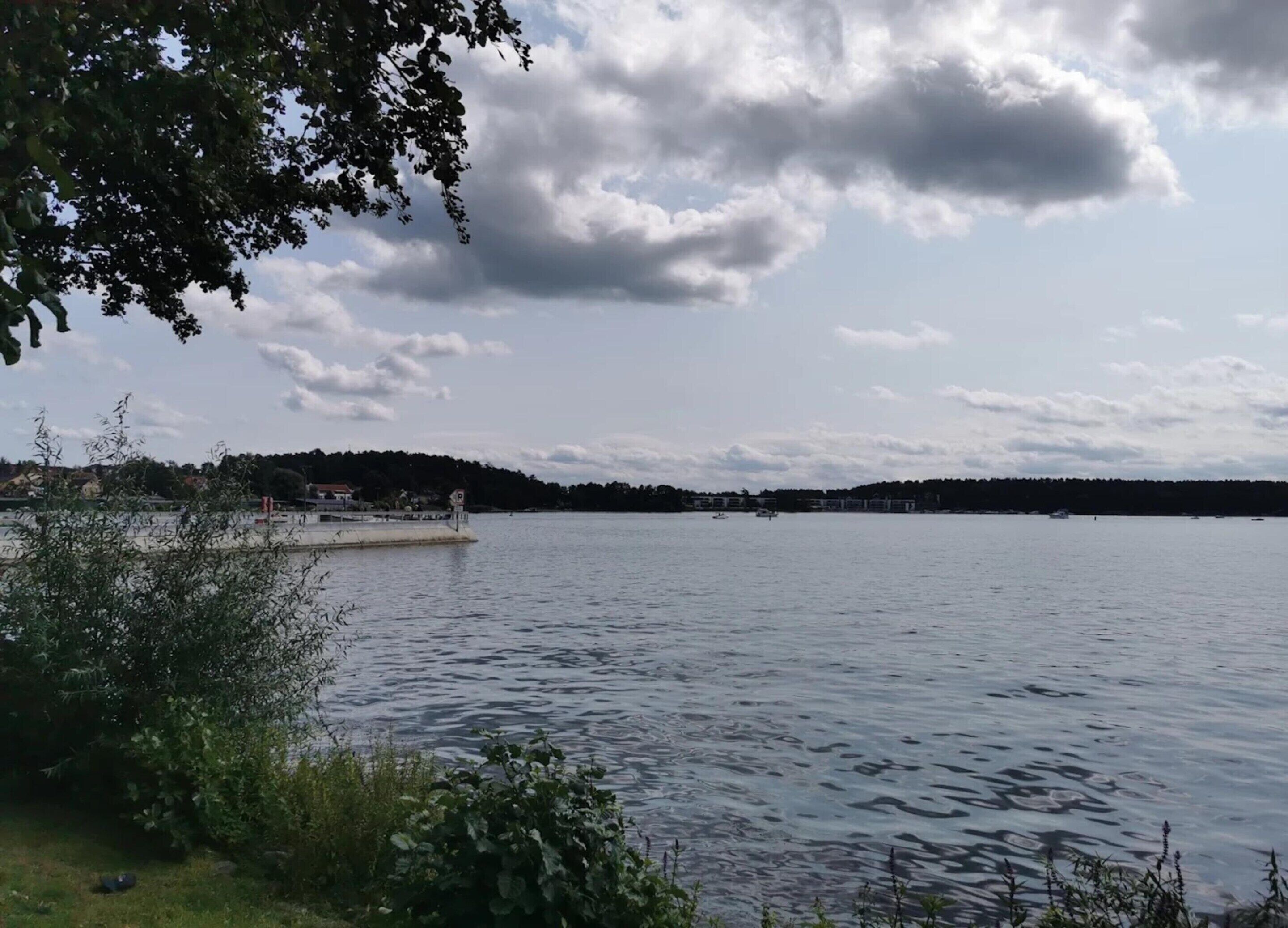 A calm lake with a dock and a small building, surrounded by greenery and a cloudy sky.