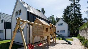 Une structure de jeux en bois, avec un toboggan et des balançoires, installée dans un espace sablonneux, avec des maisons en arrière-plan.