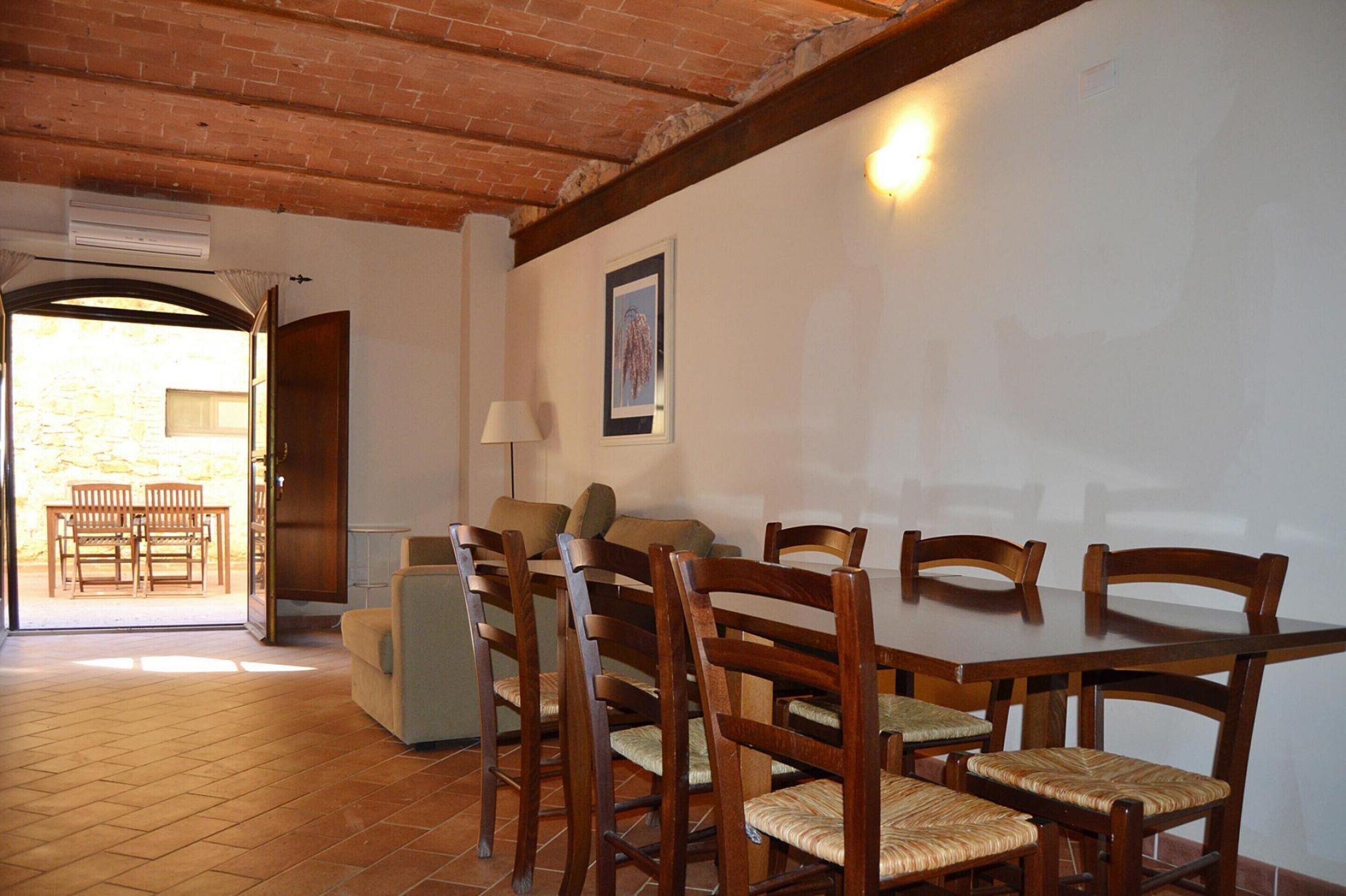 A dining area with a wooden table and chairs, a sofa, and a lamp. There is a door leading to an outdoor patio with chairs.