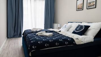 Un lit bien fait, recouvert d’une couvre-lit bleu marine ornée de motifs d’ancres, avec des oreillers blancs et un coussin décoratif portant un logo circulaire.