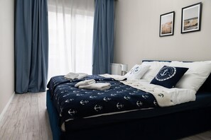 A neatly made bed with a navy blue bedspread featuring anchor patterns, white pillows, and a decorative pillow with a circular logo. - Apartment With air Conditioning, Pool, Sauna (Dziwnów)
