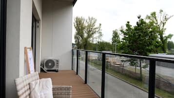 Un balcón con una silla de mimbre y un cojín, un aire acondicionado instalado en la pared y vista a una calle con árboles y edificios.