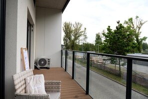Un balcon avec un fauteuil en osier et un coussin, un climatiseur fixé au mur, et une vue sur une rue bordée d’arbres et de bâtiments.