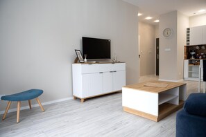 Un salon moderne avec un téléviseur à écran plat, une table basse, un banc bleu et un coin cuisine à l’arrière-plan.