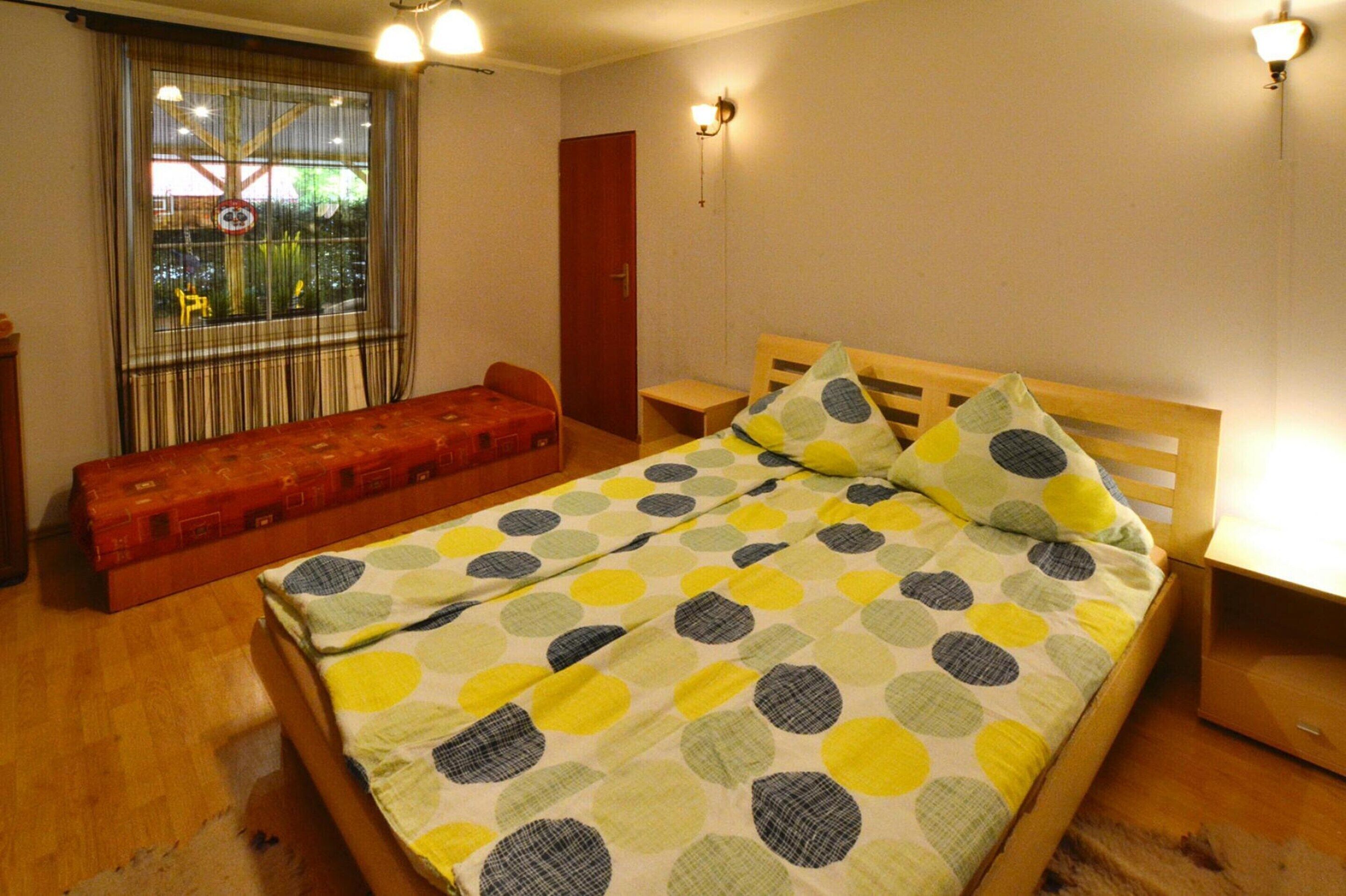A single bed with a polka dot bedspread, a wooden bench, a red door, and a window with curtains.
