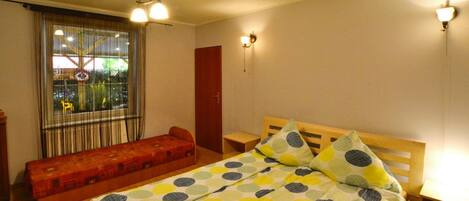 A single bed with a polka dot bedspread, a wooden bench, a red door, and a window with curtains.