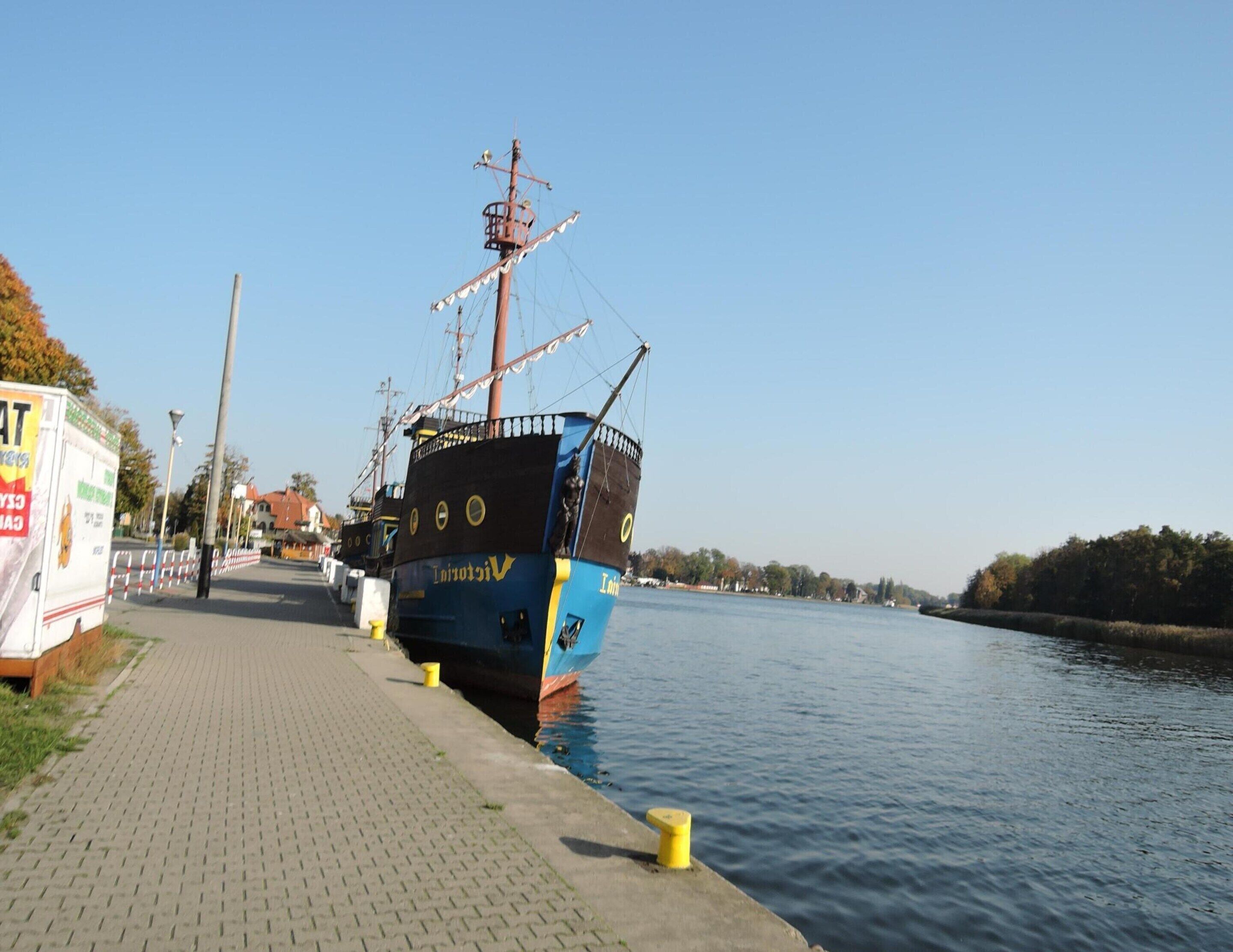 A historic ship docked at a canal with a paved walkway beside it.