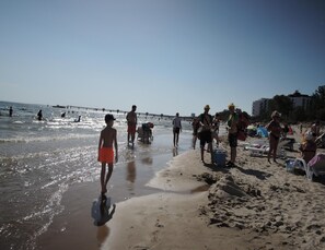 Escena de playa con gente caminando por la orilla, algunos cargando equipo de playa, y un muelle al fondo.