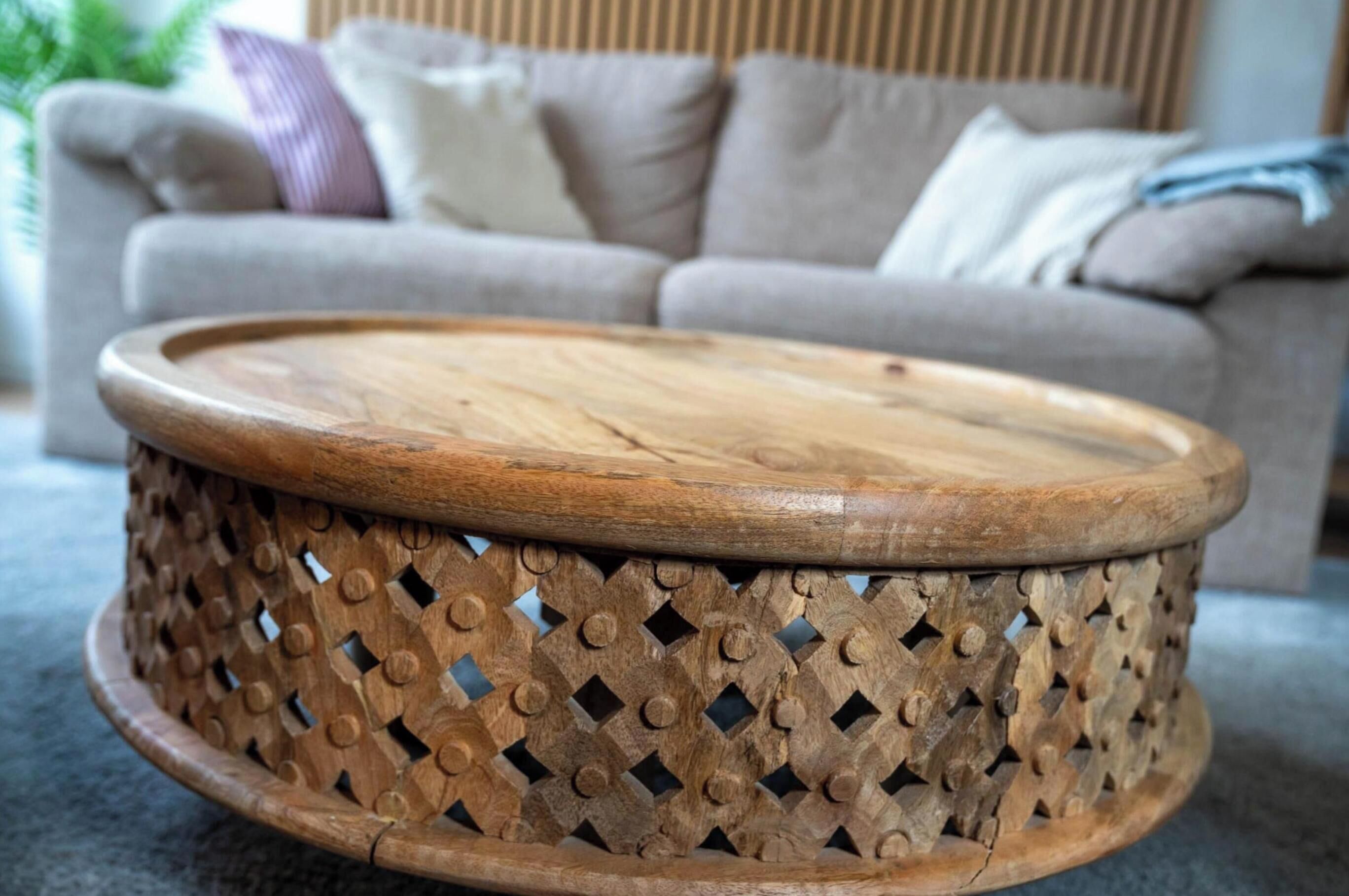 A round wooden coffee table with a lattice design, placed in a living room with a grey sofa and cushions in the background.