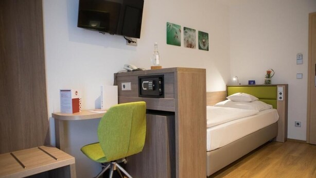 A compact hotel room with a bed, a desk, a green chair, a microwave, a television, and a small table with a lamp. - Wolfsburg Sandkamp Hotel by Belvilla (Wolfsburg)