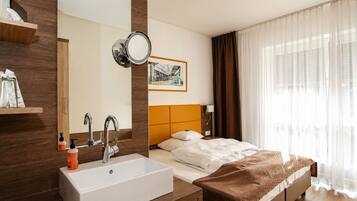 A hotel room with a large mirror, a white sink, a bed with a brown headboard, and a window with sheer curtains.