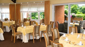 A restaurant with multiple tables and chairs, large windows with curtains, and a view of greenery outside.