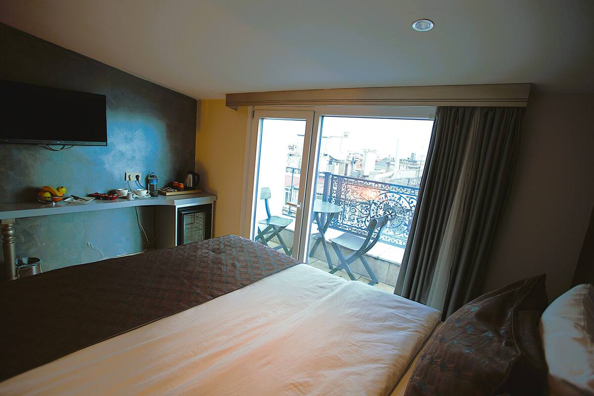 A hotel room with a bed, a desk with a flat-screen TV, a window with a view of the city, and a balcony with chairs.