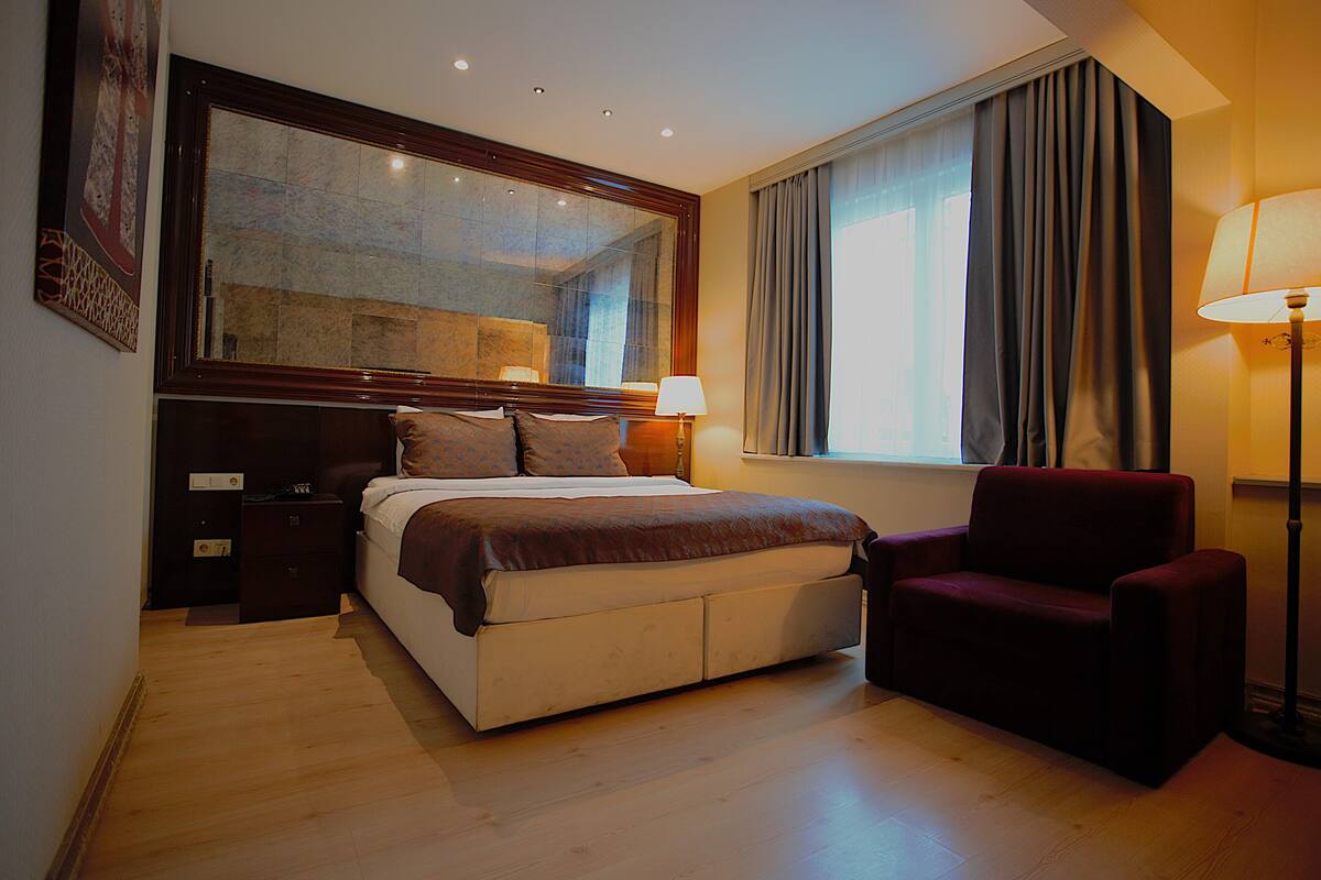 A hotel room with a large bed, a chair, a nightstand, a lamp, and a window with curtains.