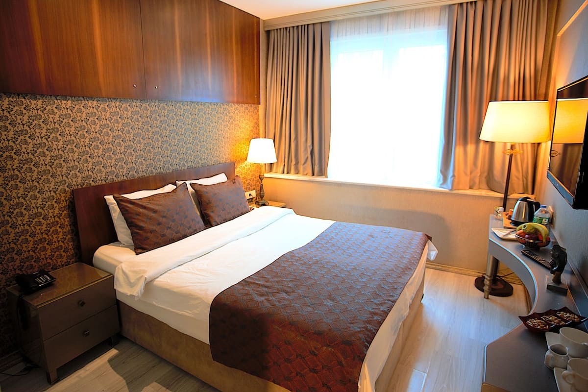 A hotel room with a large bed, two bedside tables, a nightstand, a desk with a lamp, and a window with curtains.