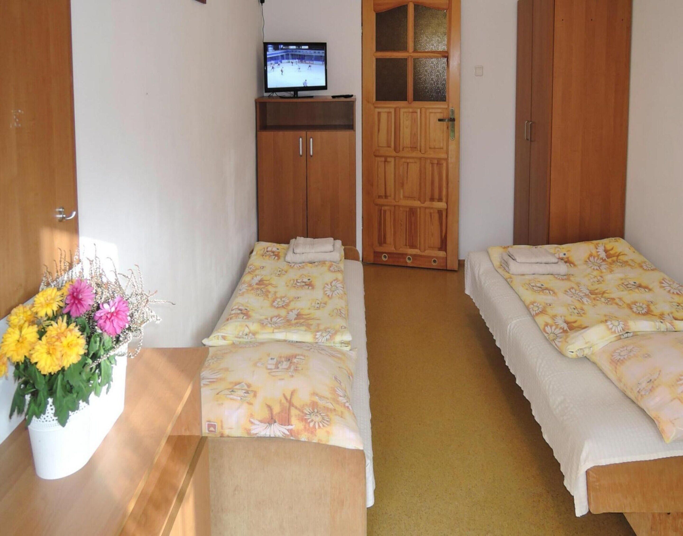 A room with two single beds, a wooden wardrobe, a television on a stand, and a vase with flowers.