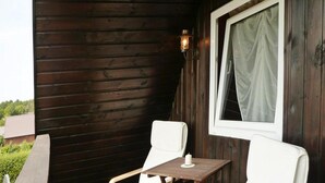A wooden deck with two white chairs and a small table, a window with a curtain, and a wooden wall. - Cottage in Koscierzyna With Lake Nearby (Koscierzyna)