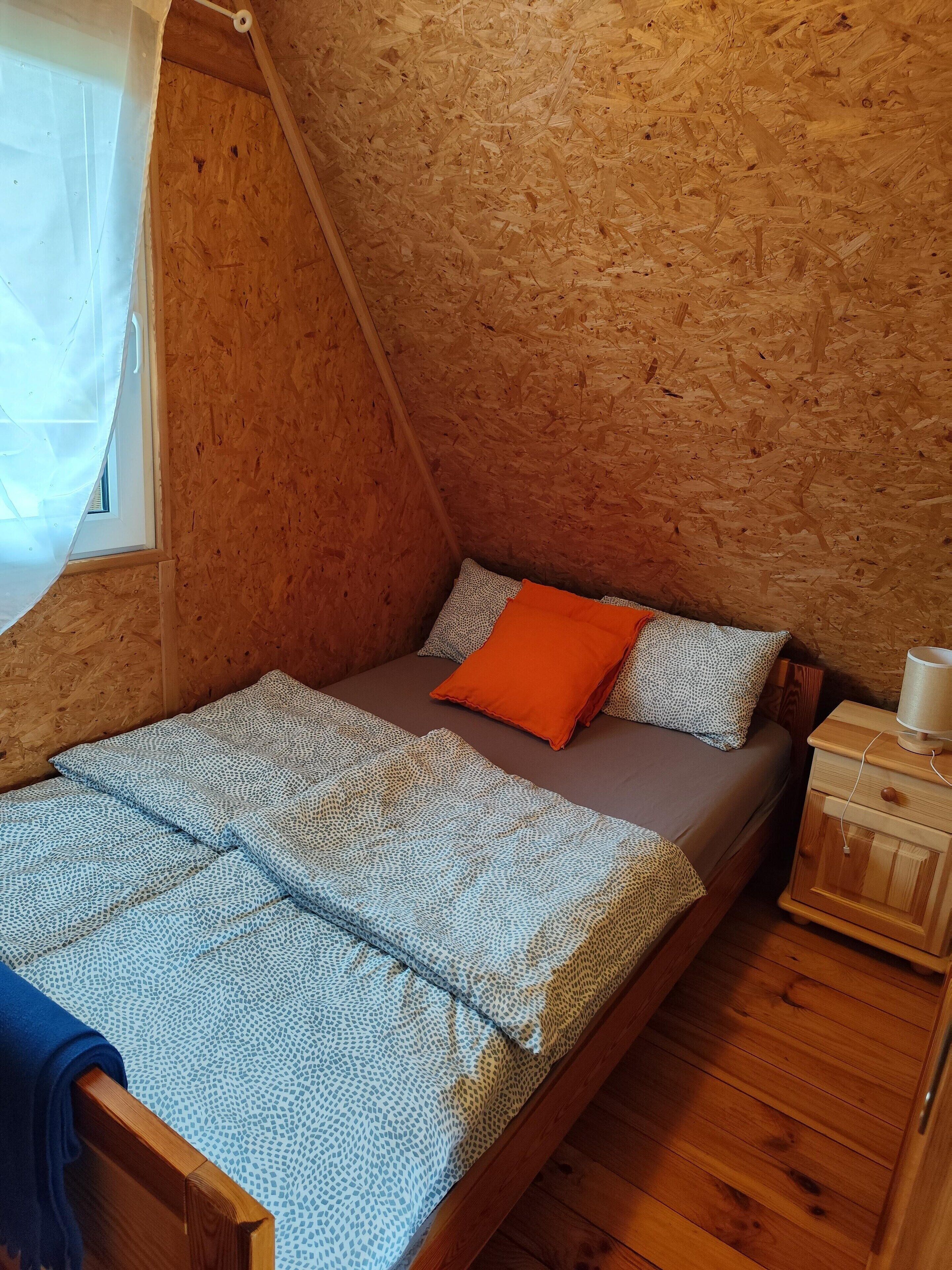Une petite pièce chaleureuse avec un plafond en pente, un lit en bois avec une literie à motifs, un oreiller orange et une table de chevet avec une lampe.