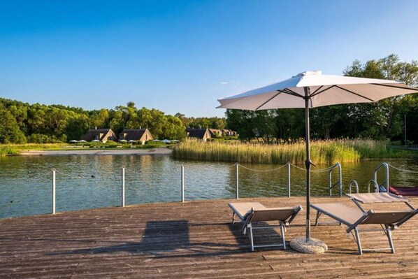 A wooden deck with lounge chairs and a large umbrella, overlooking a calm lake with houses in the background. - Appartement am Plauer See in Ganzlin (Ganzlin)