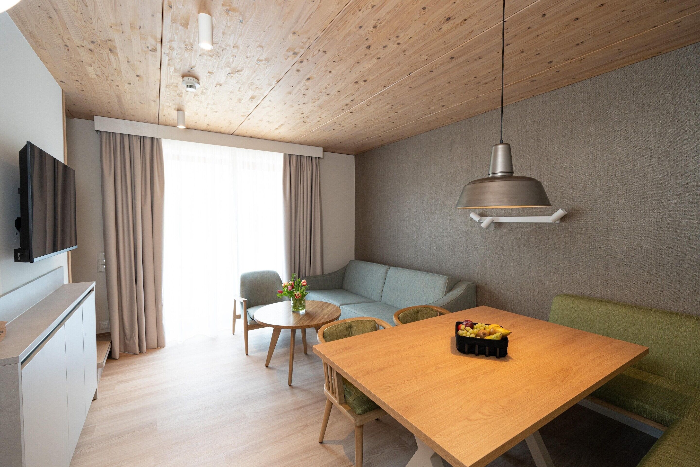 A modern living room with a wooden ceiling, a grey sofa, a wooden dining table with fruit, and a TV mounted on the wall.