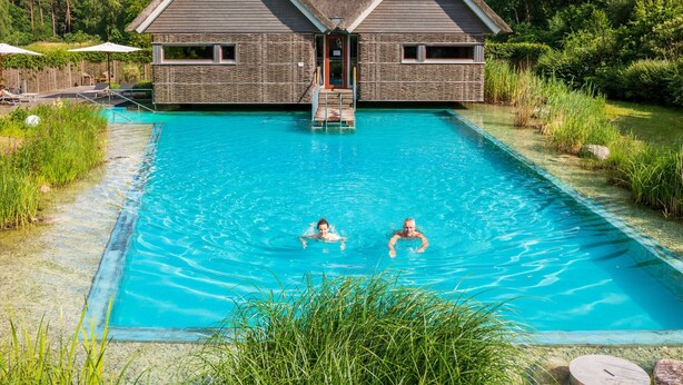 Ein Schwimmbecken mit zwei Personen im Wasser, umgeben von einem Gebäude mit einem Strohdach und Steinmauern.
