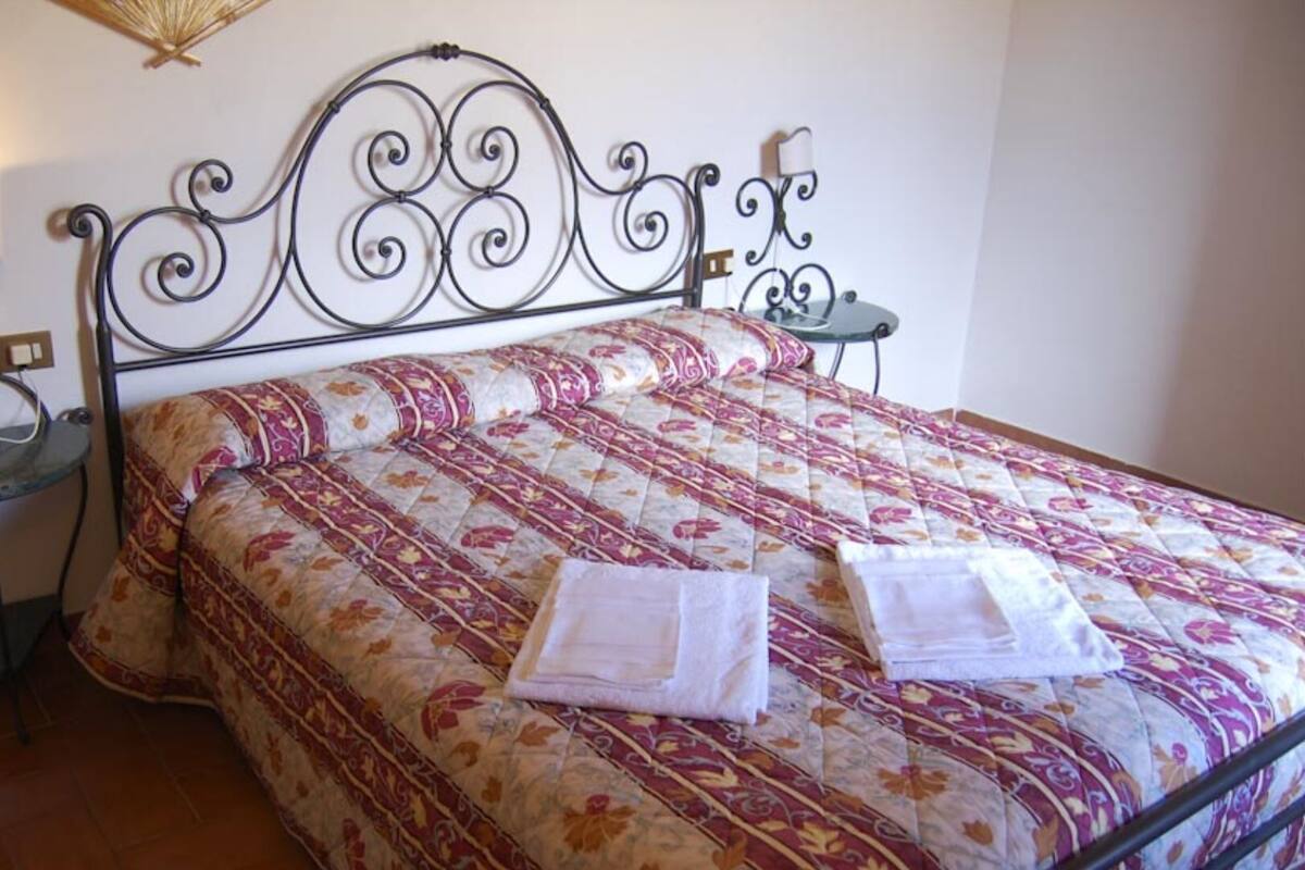 Una cama bien hecha con colcha de motivos florales y dos toallas dobladas. Hay dos mesitas de noche con lámparas a cada lado de la cama.