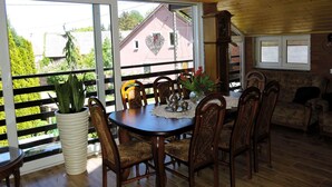 A dining area with a wooden table and chairs, a potted plant, and a view of a house through large windows.