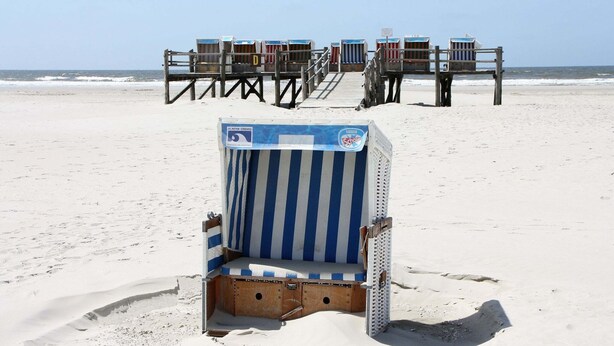 Una silla de playa con cojín azul y blanco a rayas, ubicada en una playa arenosa con huellas a su alrededor.