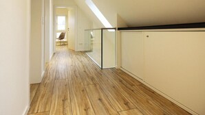 Un estrecho pasillo con suelo de madera, barandilla de cristal y una puerta que da a otra habitación.
