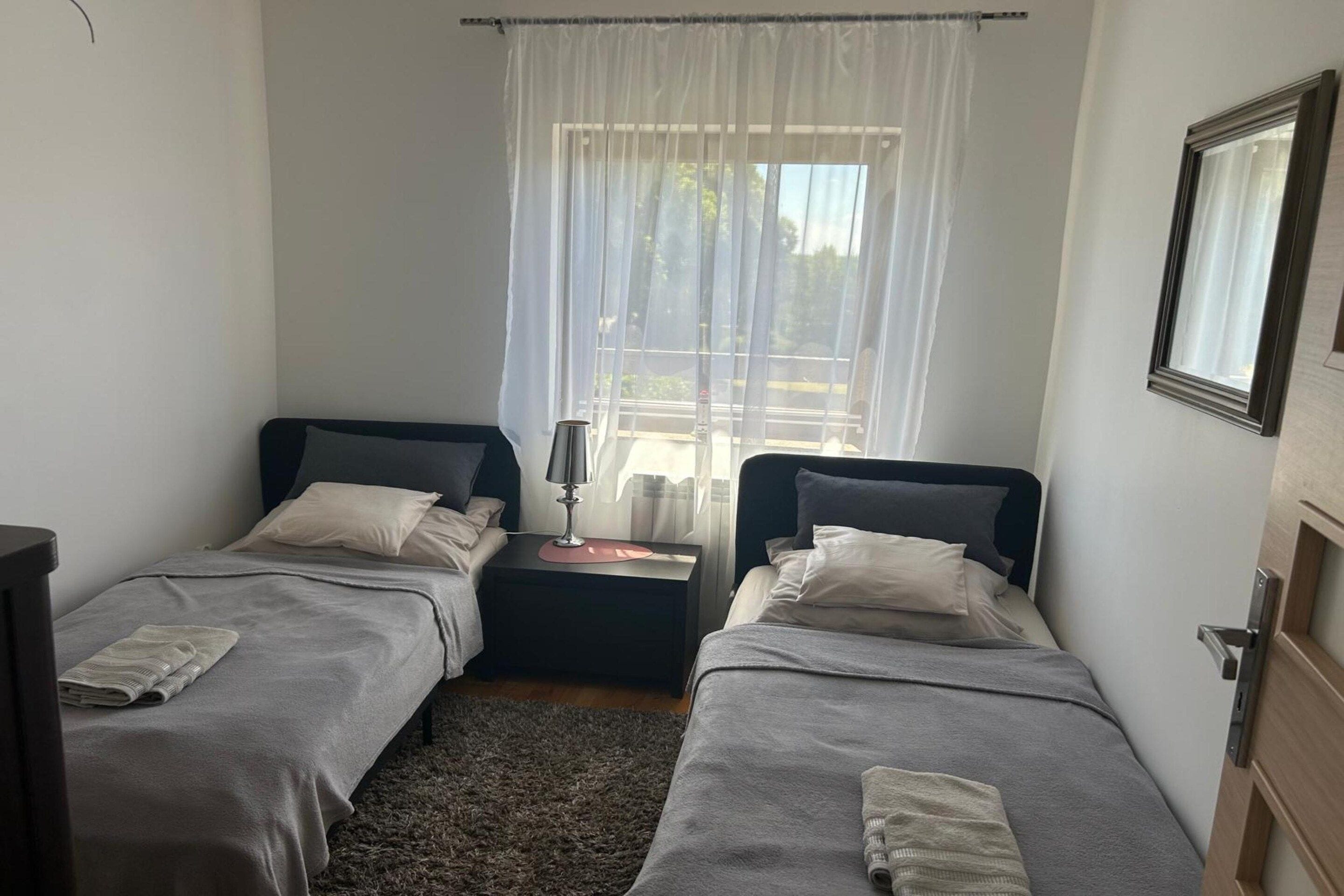 A small, single-room accommodation with two beds, a bedside table with a lamp, a window with sheer curtains, and a door with a mirror.