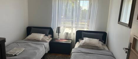 Un petit logement composé d’une seule pièce, comprenant deux lits, une table de chevet avec une lampe, une fenêtre avec des rideaux légers et une porte dotée d’un miroir.