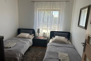 Ett litet, enkelt rum med två sängar, ett nattduksbord med en lampa, ett fönster med tunna gardiner och en dörr med en spegel.
