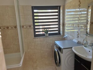 Ein Badezimmer mit Dusche, Waschmaschine, Spüle und einem Fenster mit Jalousien.
