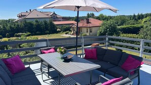 Un balcon doté d’une table, de chaises et d’un canapé, offrant une vue sur un quartier résidentiel avec ses maisons et sa verdure.
