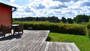 Een houten terras met tuinmeubilair, een rood gebouw en uitzicht op een groen veld en bomen onder een gedeeltelijk bewolkte lucht.