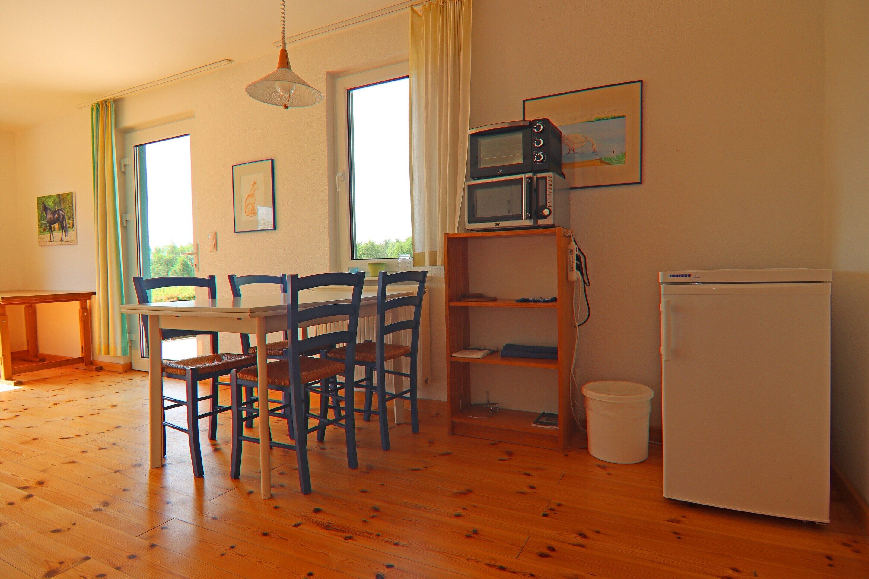 Een eethoek met een houten tafel en stoelen, een witte koelkast, een magnetron op een plank, en een raam met gordijnen.