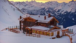 Eine Berglodge mit Steinaußenwand und Holzakzenten, umgeben von schneebedeckten Gipfeln und einem Abendhimmel.